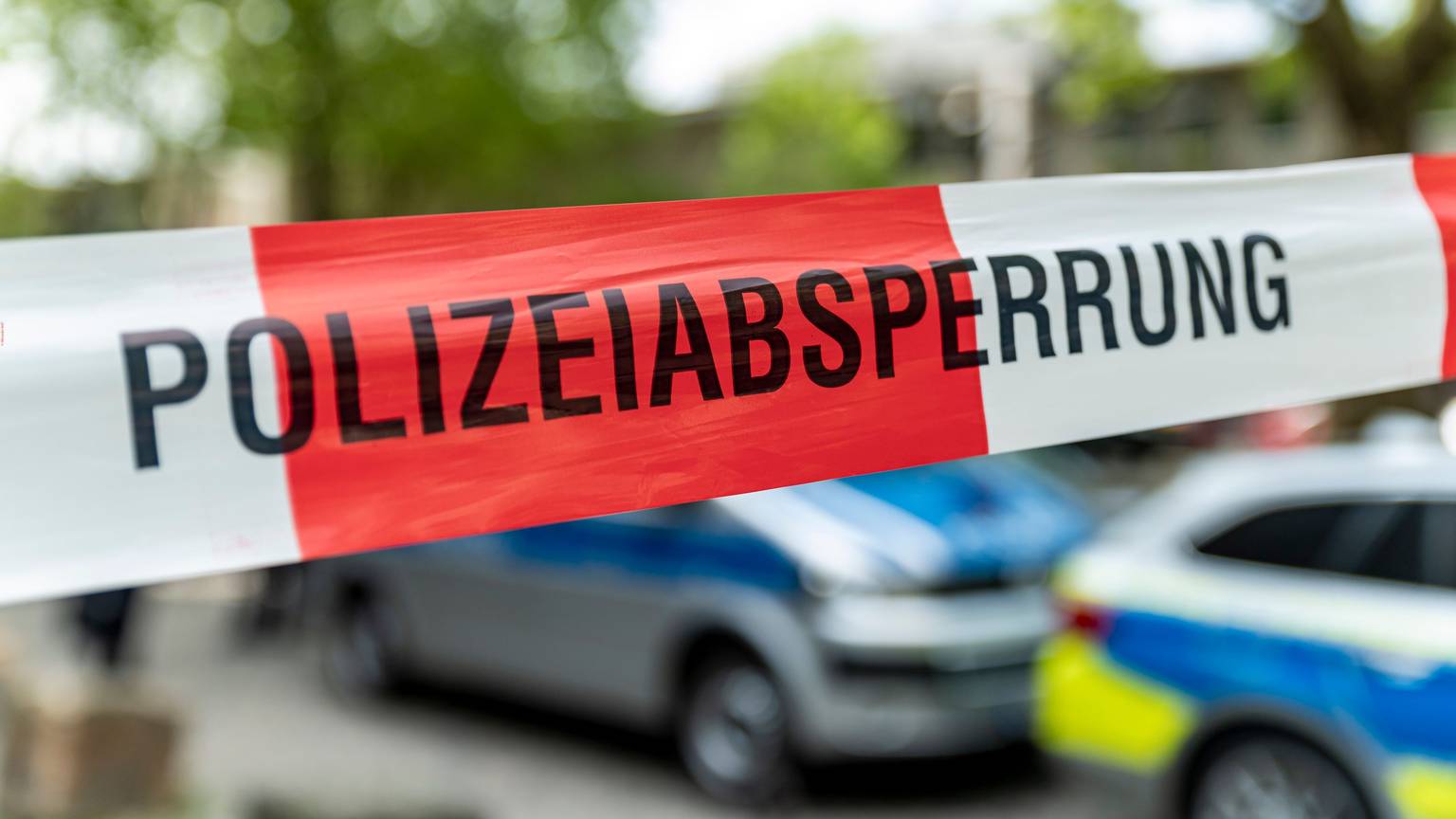 Auf rot-weißem Flatterband steht das Wort „Polizeiabsperrung“. Dahinter stehen einige Polizeiwagen.