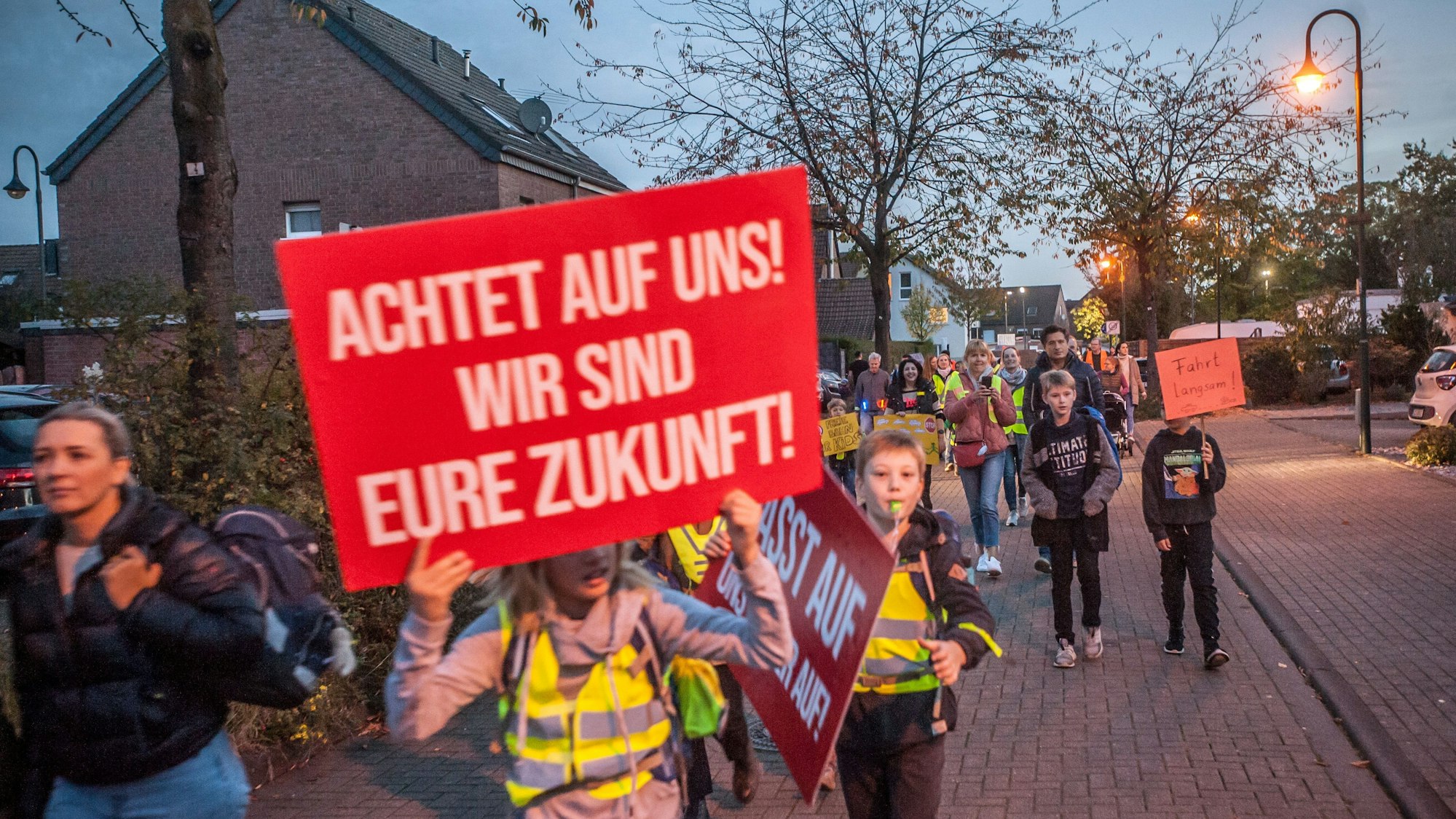 Kinder mit Plakaten und in elterlicher Begleitung protestieren in der Dämmerung auf der Hitdorfer Ringstraße für mehr Sicherheit