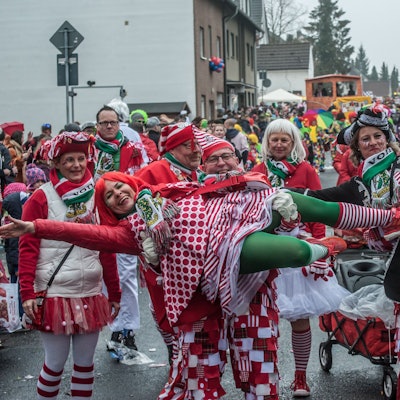 Verkleidete Männer im Karnevalszug heben zwei verkleidete Frauen elegant hoch.