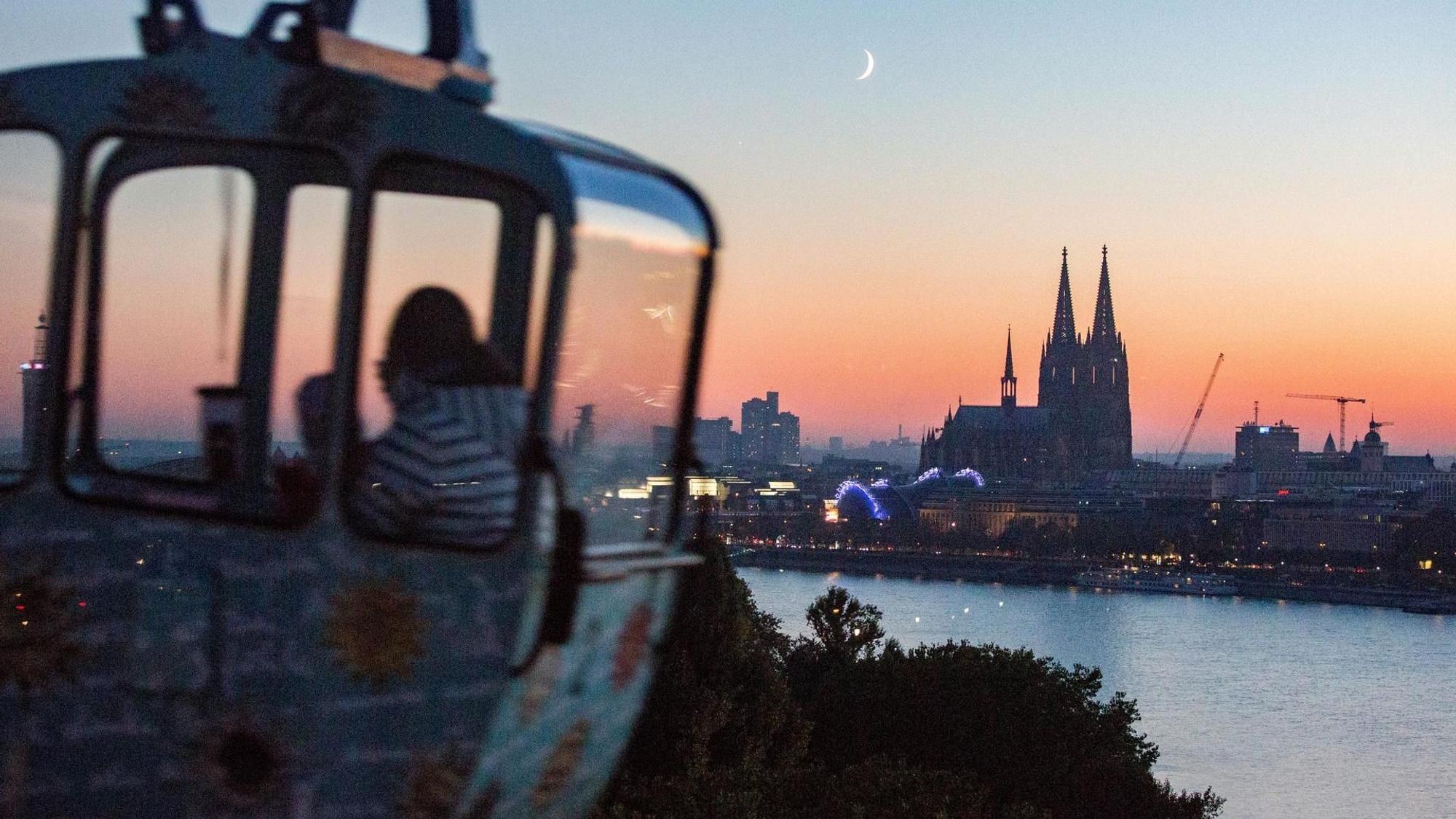 Eine Gondel der Seilbahn ist vor dem Sonnenuntergang am Kölner Rheinufer zu sehen, im Mittelpunkt der Dom.