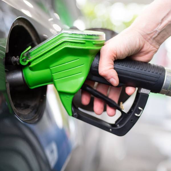 Eine Frau hält an einer Tankstelle an einer Zapfsäule eine Zapfpistole in der Hand und betankt ein Auto.