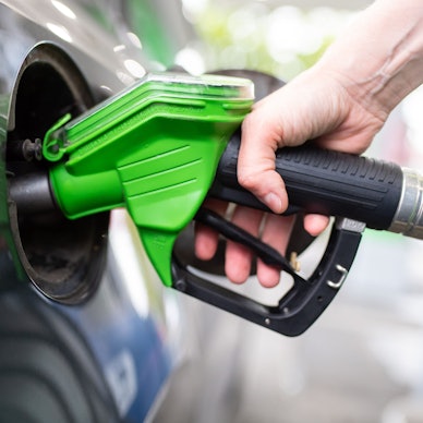 Eine Frau hält an einer Tankstelle an einer Zapfsäule eine Zapfpistole in der Hand und betankt ein Auto.