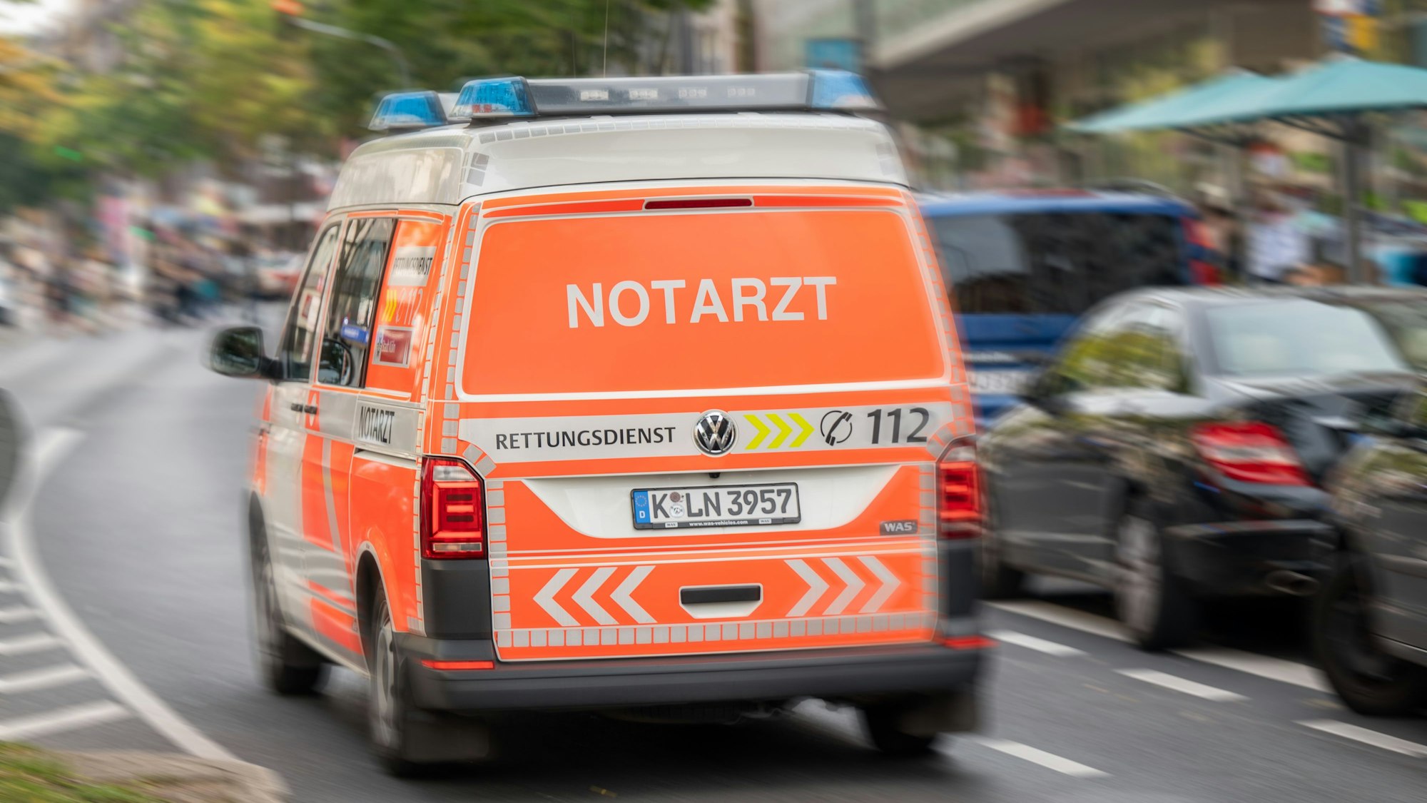 Ein Notarztwagen ist auf einer Straße unterwegs. Der Notarztwagen fährt an parkenden Autos am rechten Straßenrand vorbei.