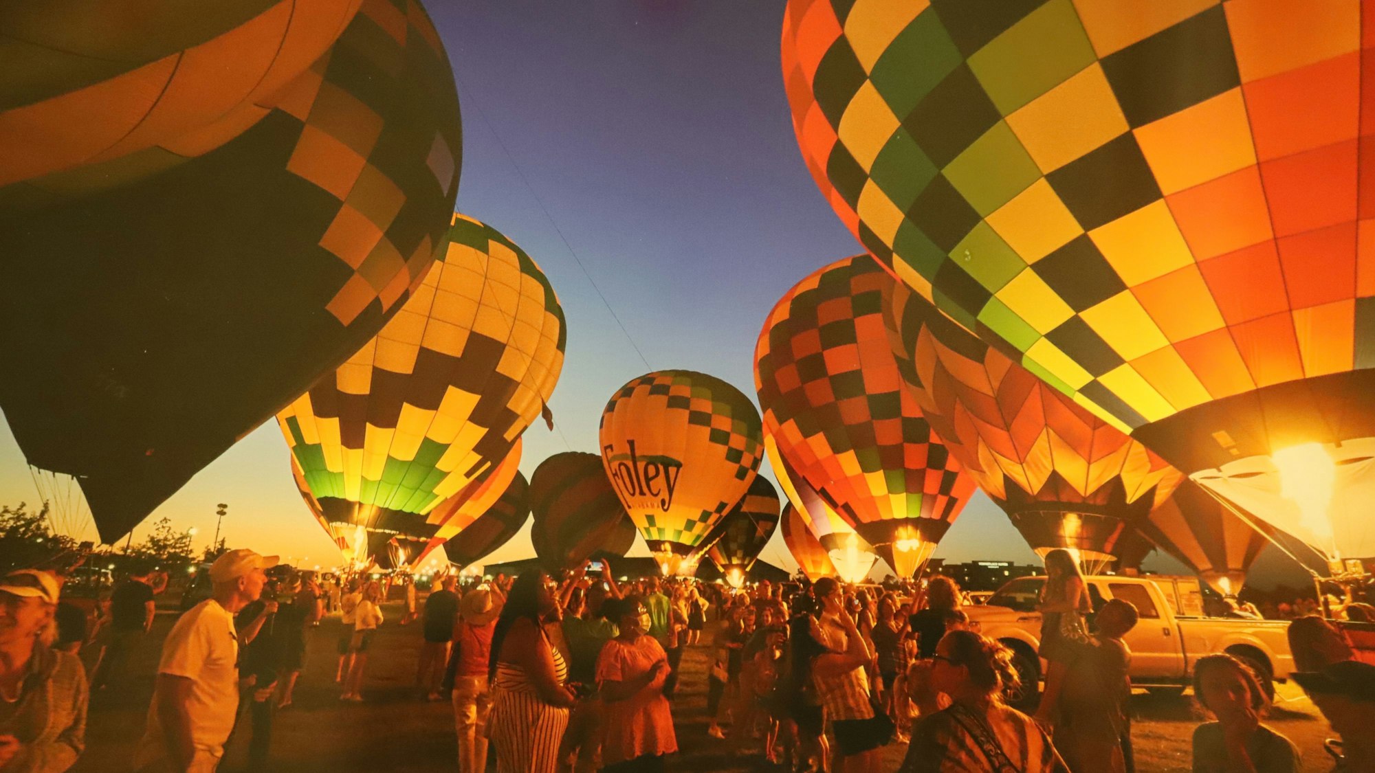 Ein Dutzend Heißluftballons stehen am Boden und glühen. Viele Menschen stehen um die Luftballons verteilt. Auf einem Ballon ist „Foley“ zu lesen.