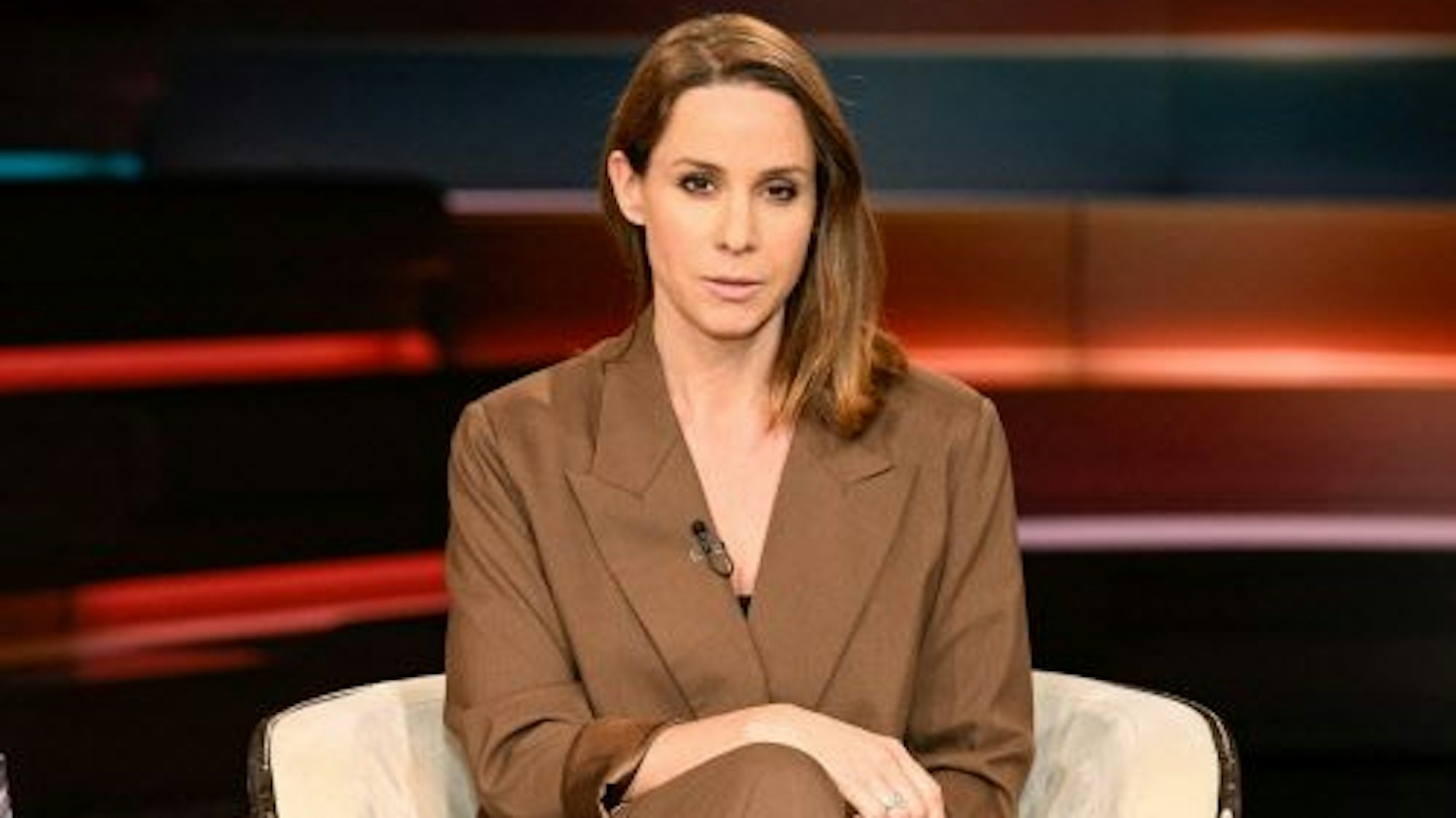 Natalie Amiri sitzt im Studio auf einem Sessel. Ihre Beine sind überschlagen und ihre Hände liegen auf ihrem Schoß. Sie guckt ernst in die Kamera.