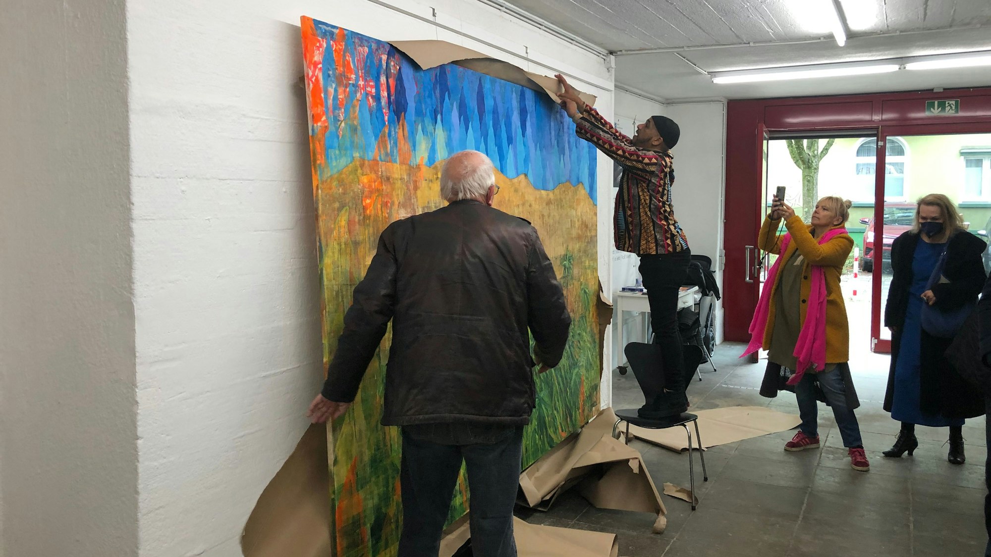 Ein Besucher der Ausstellung im Opladener Künstlerbunker reißt Packpapier auf, mit dem ein großes, buntes Kunstwerk verdeckt wurde. Andere Besucher beobachten ihn dabei.