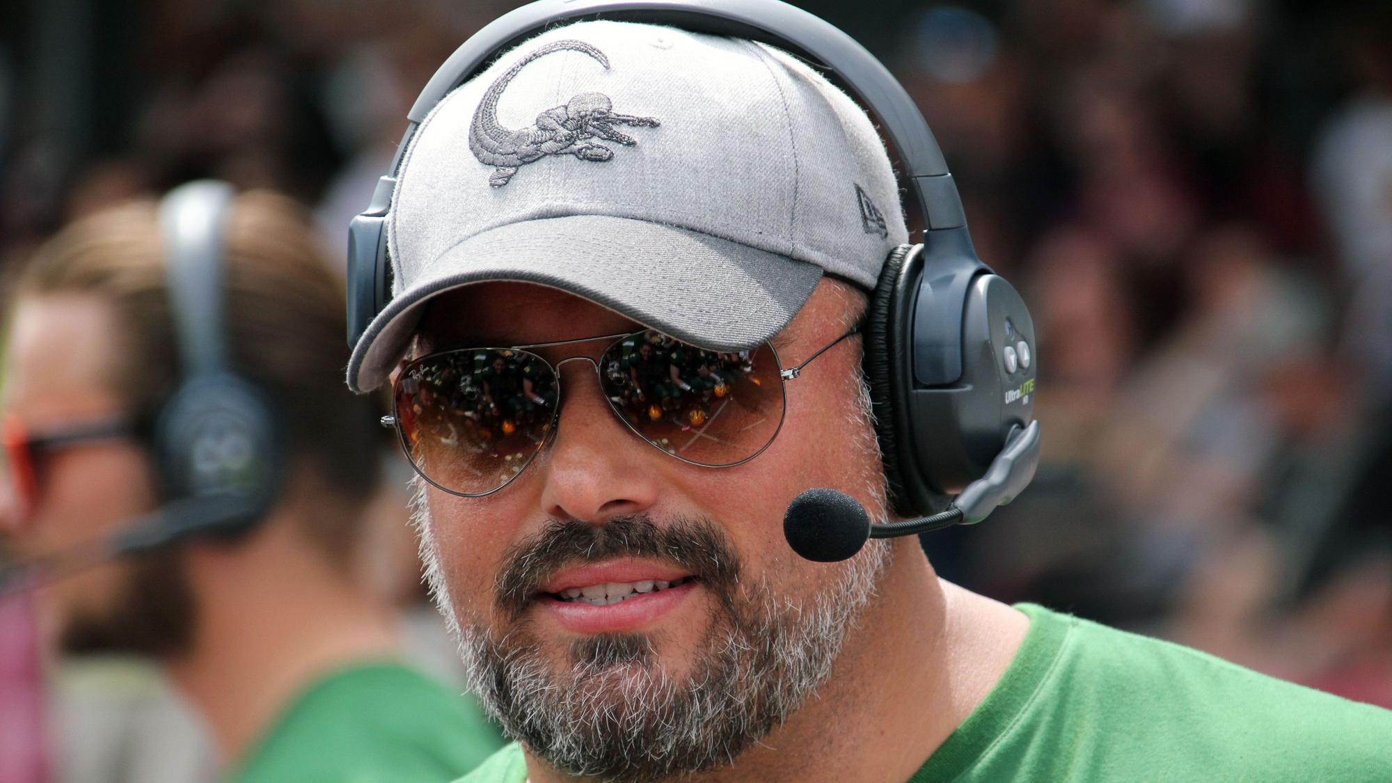Ein Football-Trainer steht mit Kappe, Kopfhörern und Sonnenbrille am Spielfeldrand.