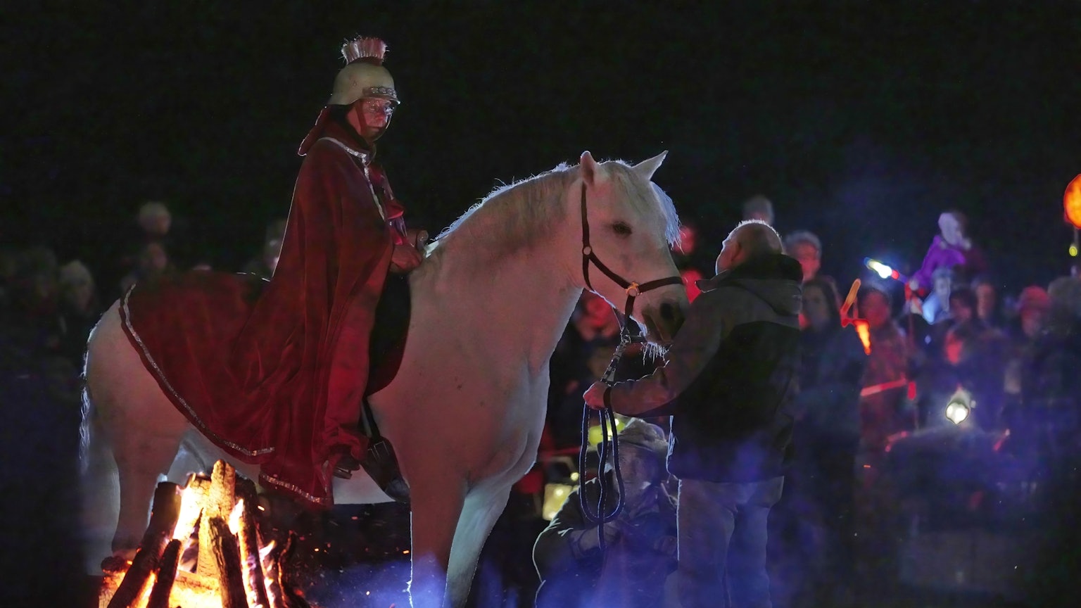 Eine Reiterin im roten Mantel und Helm auf dem Kopf sitzt auf einem Schimmel. Im Hintergrund sind Kinder zu sehen.