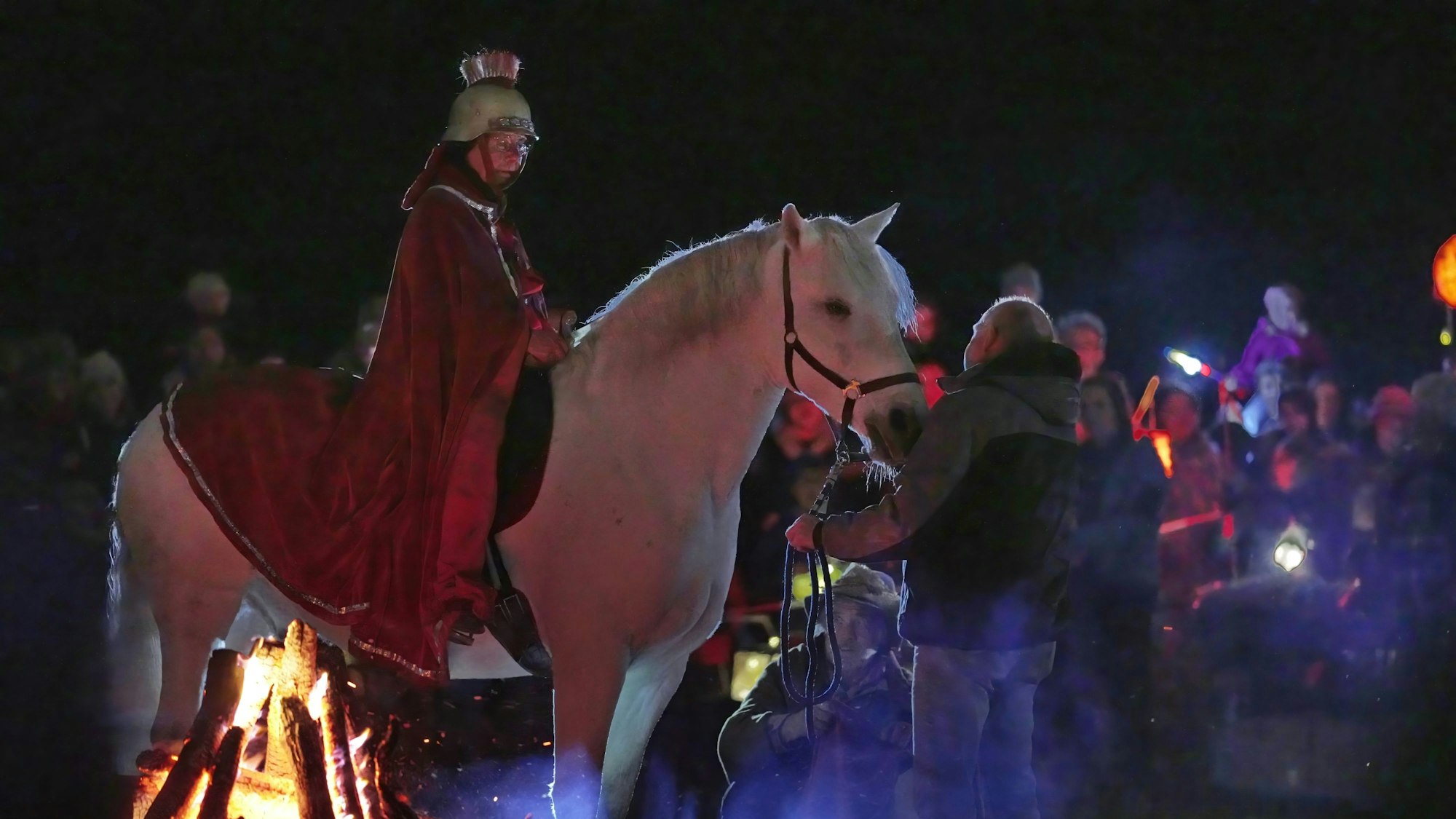 Eine Reiterin im roten Mantel und Helm auf dem Kopf sitzt auf einem Schimmel. Im Hintergrund sind Kinder zu sehen.