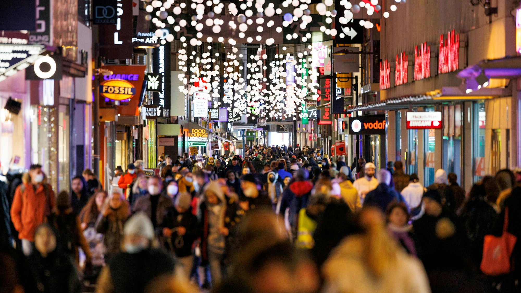 Zu sehen ist die Hohe Straße in Köln, viele Menschen drängen sich durch die Straße, der Vordergrund ist verschwommen. Es ist mit Weihnachtsdekoration geschmückt und es ist dunkel.