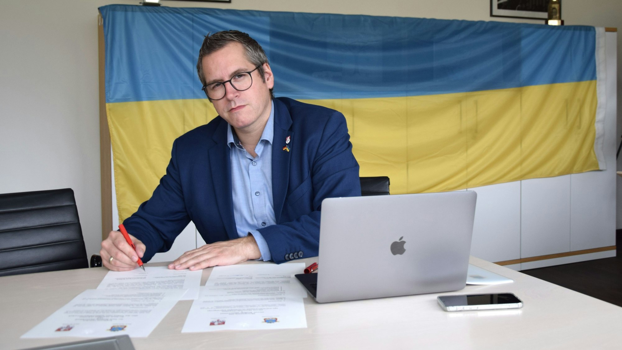 Bedburgs Bürgermeister Sascha Solbach unterzeichnet die Urkunde zur Solidaritätspartnerschaft mit Mykolajiw. Im Hintergrund ist eine Ukraine-Flagge zu sehen.