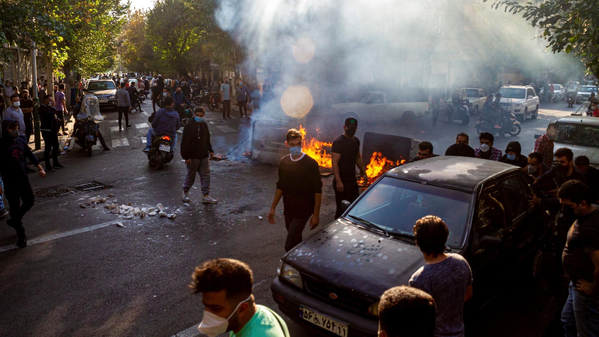 Menschen protestieren auf den Straßen in Teheran, der iranischen Hauptstadt. Zwischen den Protestierenden sind brennende Gegenstände und Rauch zu sehen.