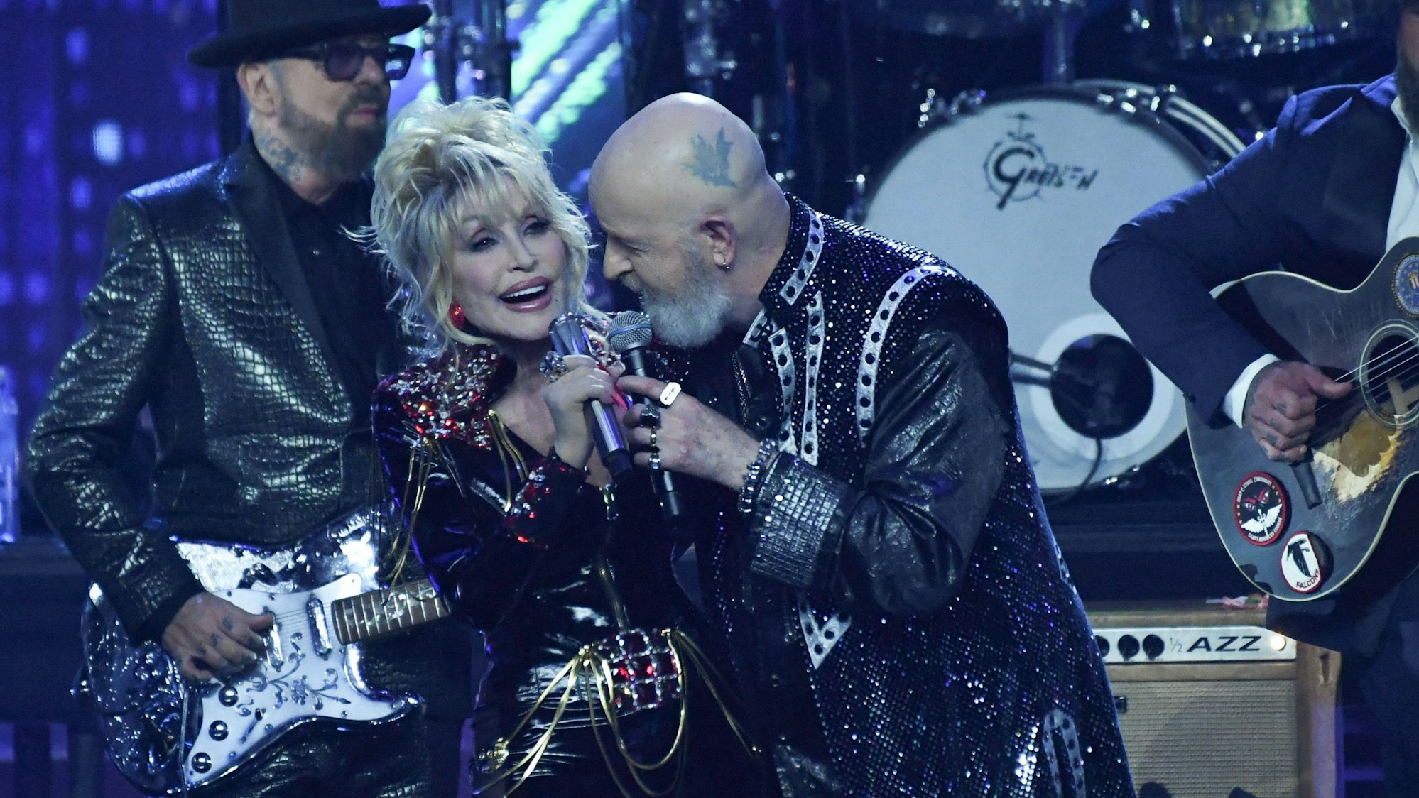 Dolly Parton Arm in Arm mit Rocksänger Rob Halford. Beide tragen Schwarz mit Glitzerelementen und silbernem Schmuck.