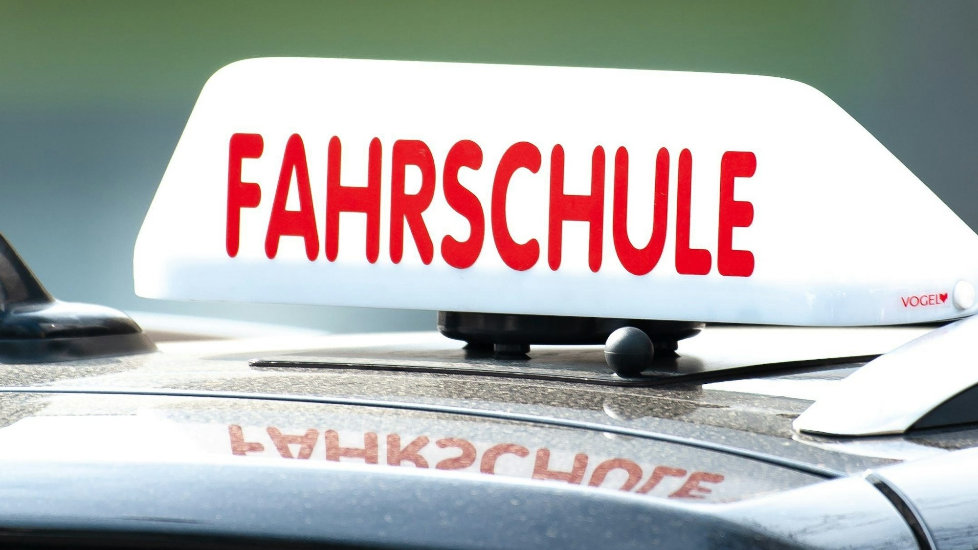 Ein weißes Fahrschul-Schild mit roter Aufschrift ist auf einem Autodach zu sehen.