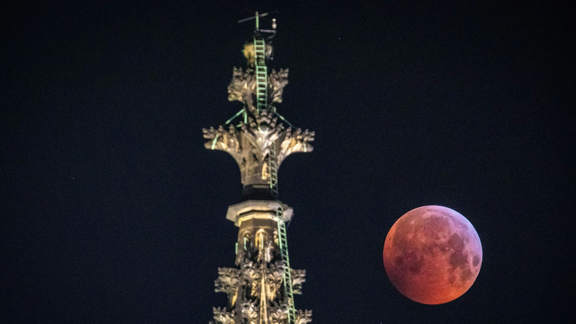 Der rot schimmernde Vollmond steht während einer totalen Mondfinsternis am Himmel neben der Kreuzblume an der Spitze des Nordturm des Kölner Dom (Archivbild).