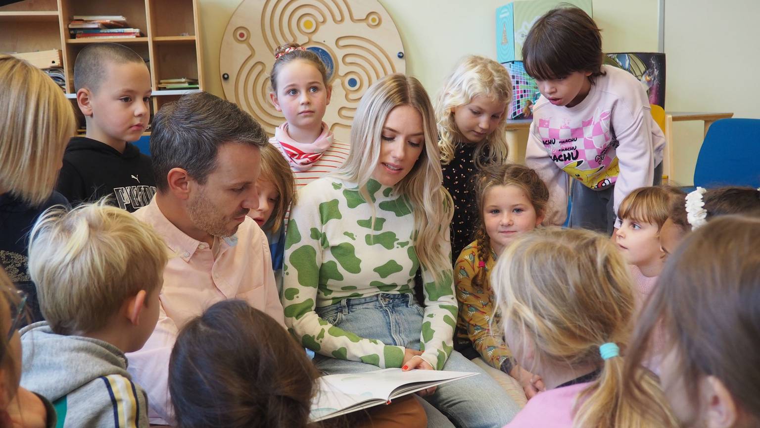Marc Dumitru und Vanessa Meisinger sitzen in einem Kreis von Grundschulkindern und lesen aus Büchern vor.