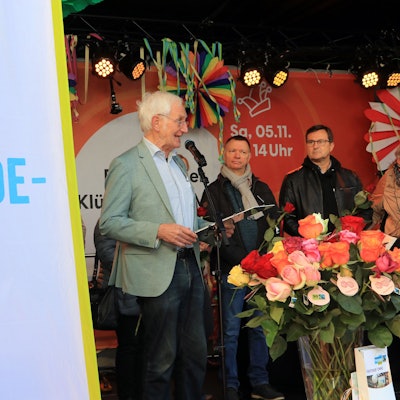 Auf einer Bühne stehen drei Männer hinter einer Flagge mit der Aufschrift „Wir sind Fair-Trade-Stadt“, unter anderem Ehrenbotschafter Manfred Holz am Mikrofon.