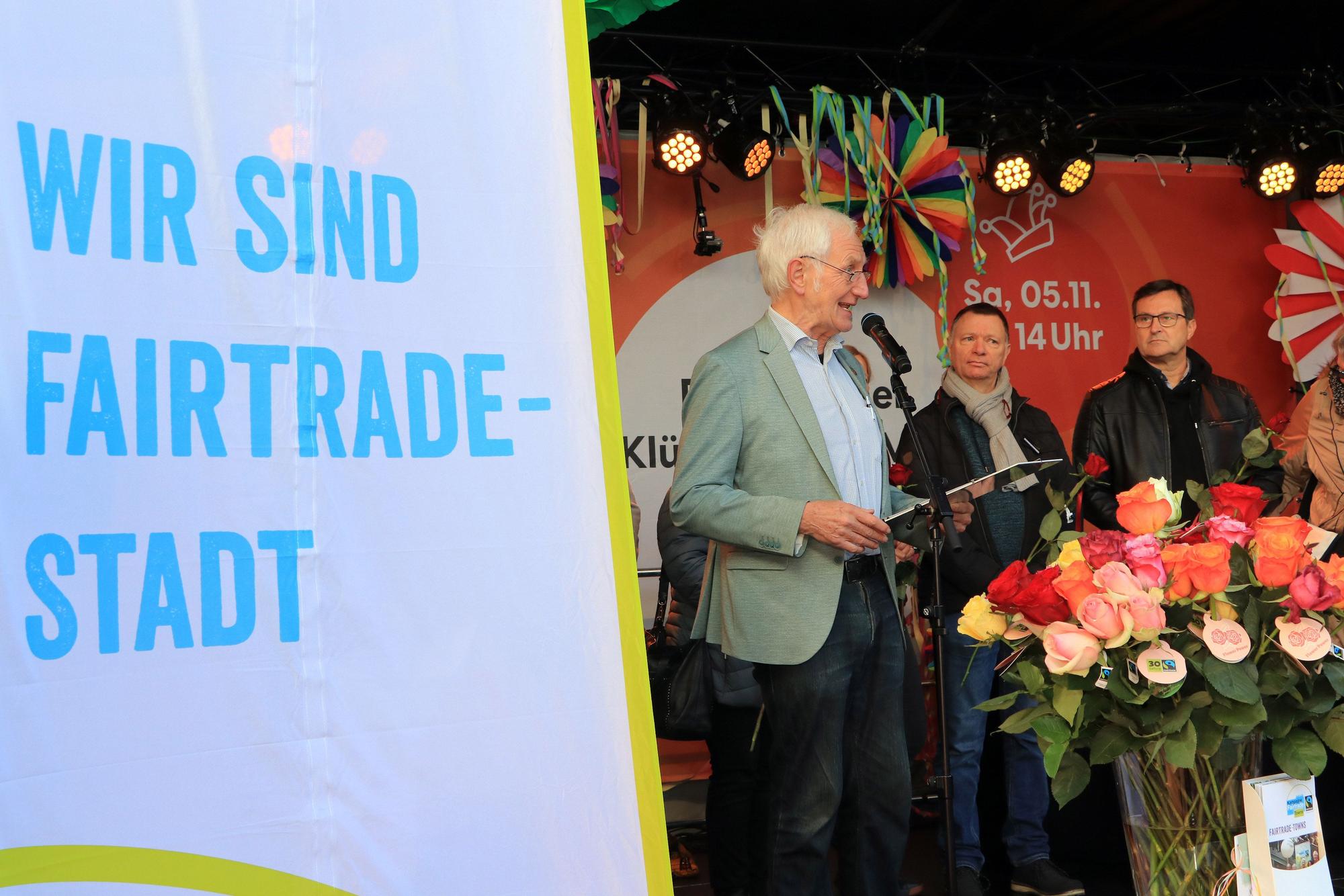 Auf einer Bühne stehen drei Männer hinter einer Flagge mit der Aufschrift „Wir sind Fair-Trade-Stadt“, unter anderem Ehrenbotschafter Manfred Holz am Mikrofon.