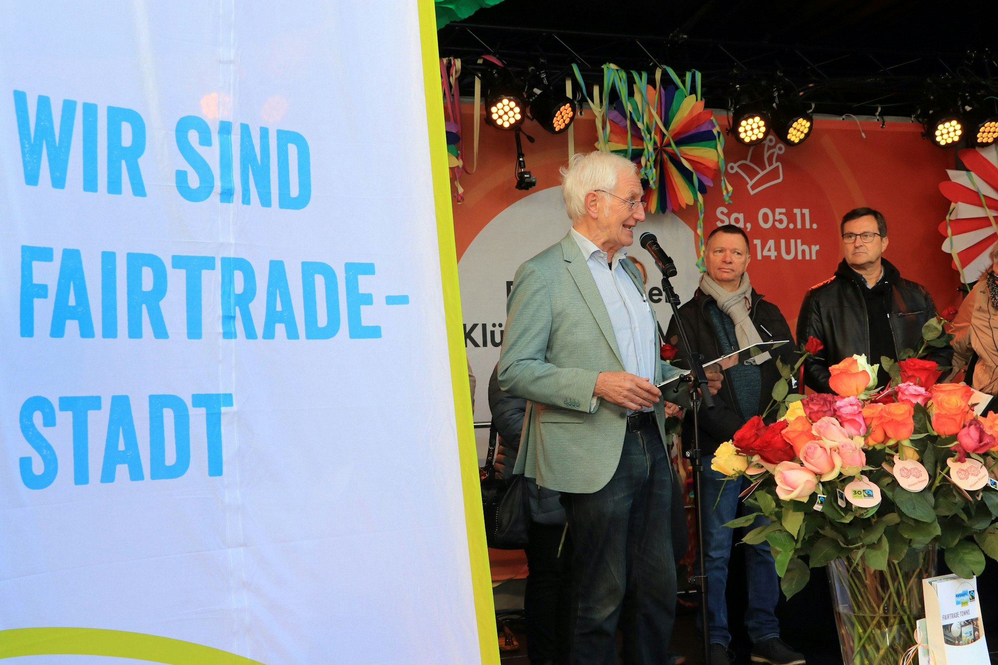 Auf einer Bühne stehen drei Männer hinter einer Flagge mit der Aufschrift „Wir sind Fair-Trade-Stadt“, unter anderem Ehrenbotschafter Manfred Holz am Mikrofon.