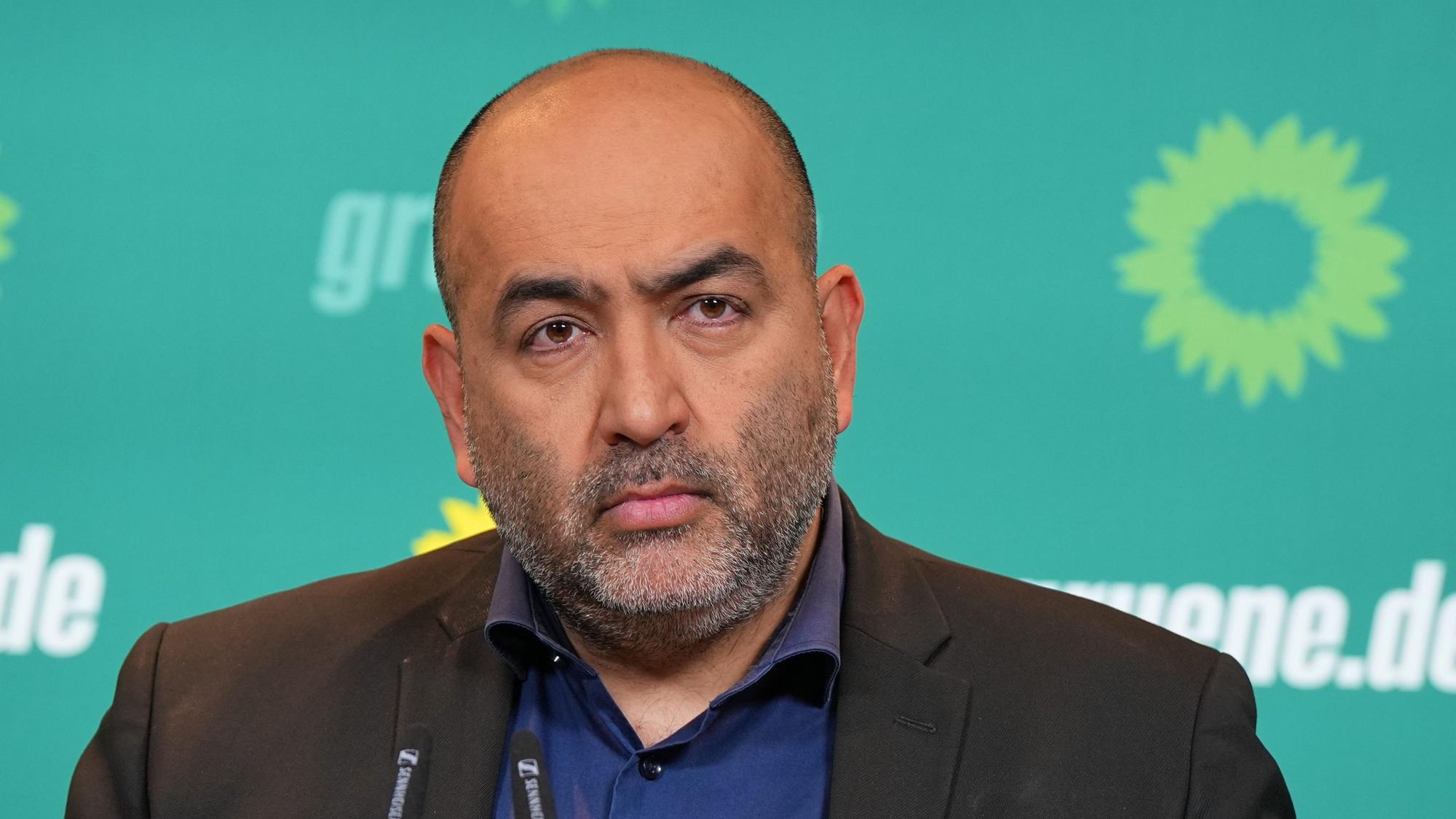 Omid Nouripour steht vor grünem Hintergrund mit Logos der Grünen drauf. Er hat eine Augenbraue hochgezogen und guckt eher traurig.