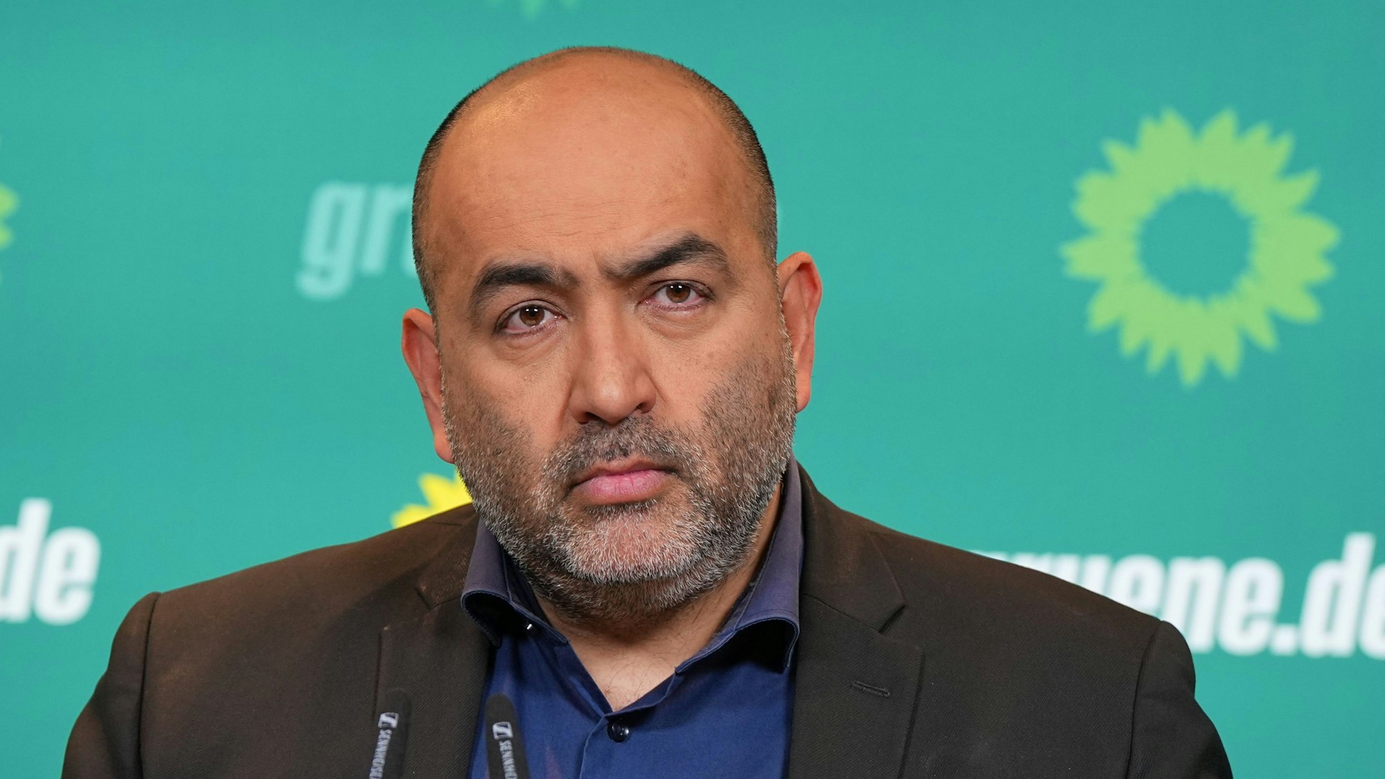 Omid Nouripour steht vor grünem Hintergrund mit Logos der Grünen drauf. Er hat eine Augenbraue hochgezogen und guckt eher traurig.