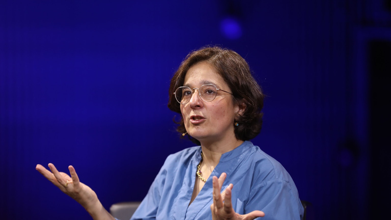 Isabel Schayani sitzt vor blauen Hintergrund und redet. Dabei gestikuliert sie mit ihren Händen.