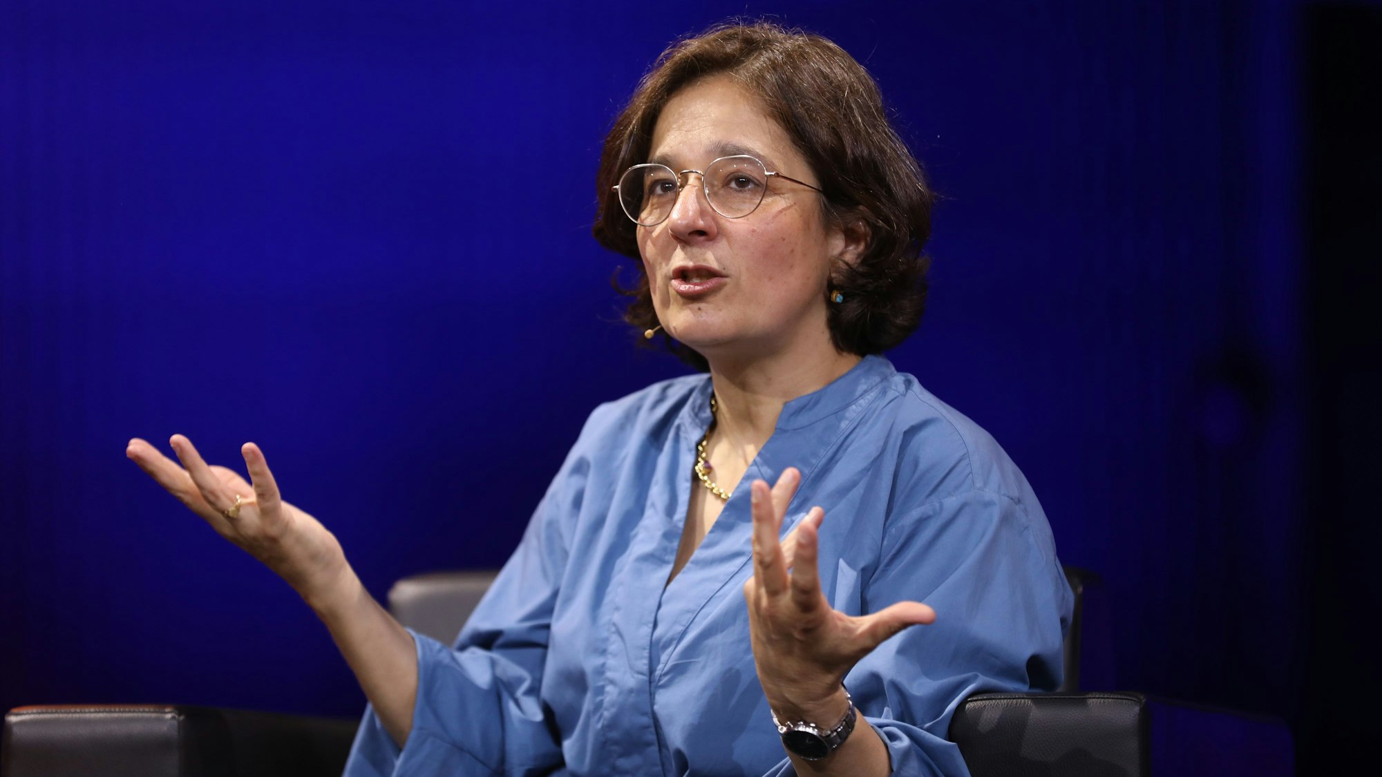 Isabel Schayani sitzt vor blauen Hintergrund und redet. Dabei gestikuliert sie mit ihren Händen.