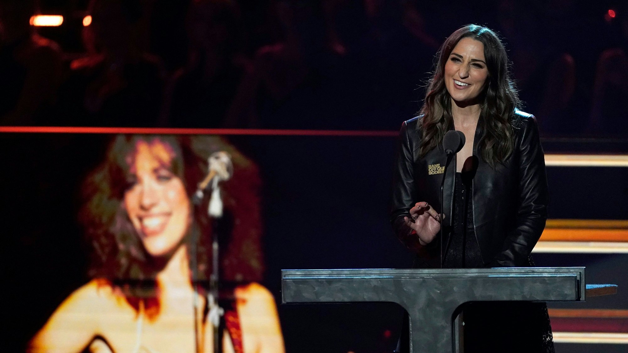Sara Bareilles in schwarzer Kleidung hält die Laudatio auf Carly Simon am Mikro. Sie steht vor einem Tisch und hebt die Hand. Sie lächelt dabei. Im Hintergrund ist ein Bild von Carly Simon zu sehen.