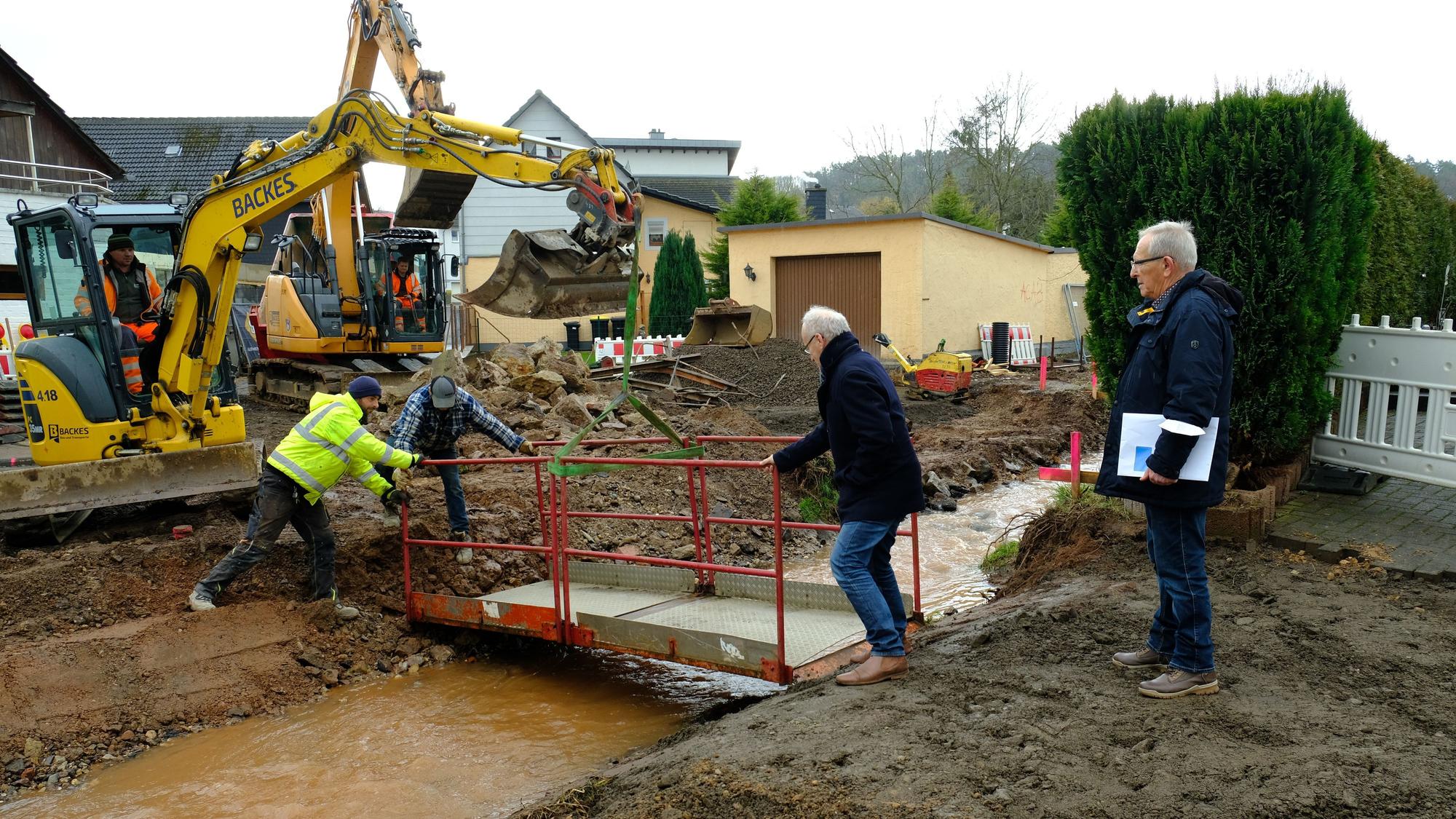 Bauarbeiter arbeiten mit einem Bagger an der Ackergasse in Kommern. Zu sehen ist auch eine provisorische Brücke, die über den Bleibach verläuft.