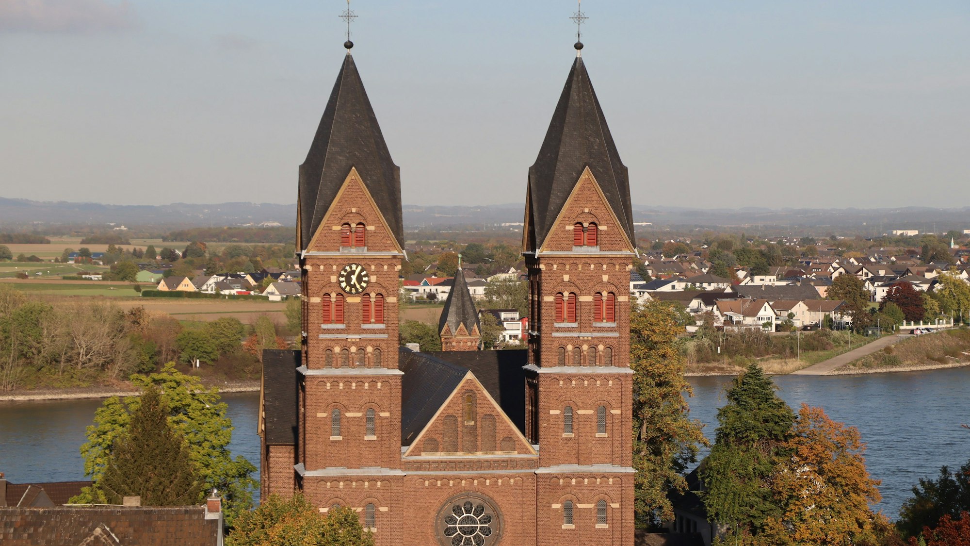 Die Kirchtürme der Pfarrkirche St. Germanus in Wesseling sind vor einem Gewässer zu sehen.