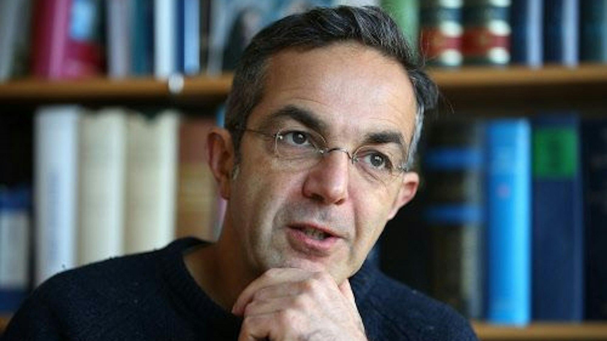 Navid Kermani sitzt vor einem Bücherregal. Er scheint zu reden und hat sein Kinn auf der Hand abgestützt.