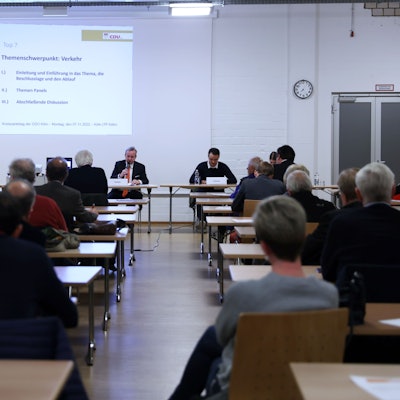 Deligierte der Kölner CDU sitzen an Tischen und blicken auf eine Präsentation des Parteichefs Bernd Petelkau.