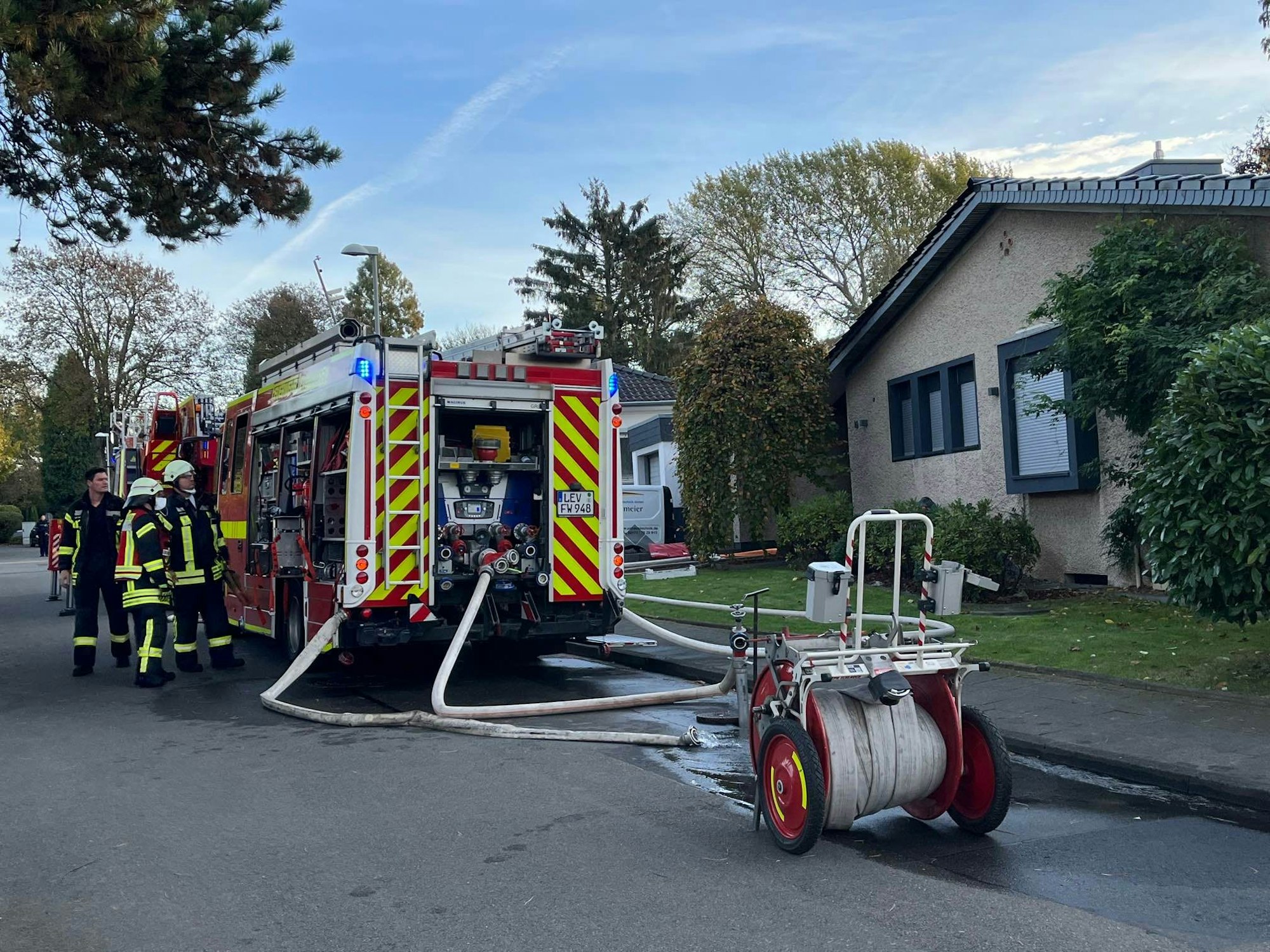 Ein Feuerwehrauto und drei Feuerwehrleute stehen auf einer Straße vor einem Haus. An dem Feuerwehrauto ist ein Schlauch angeschlossen.