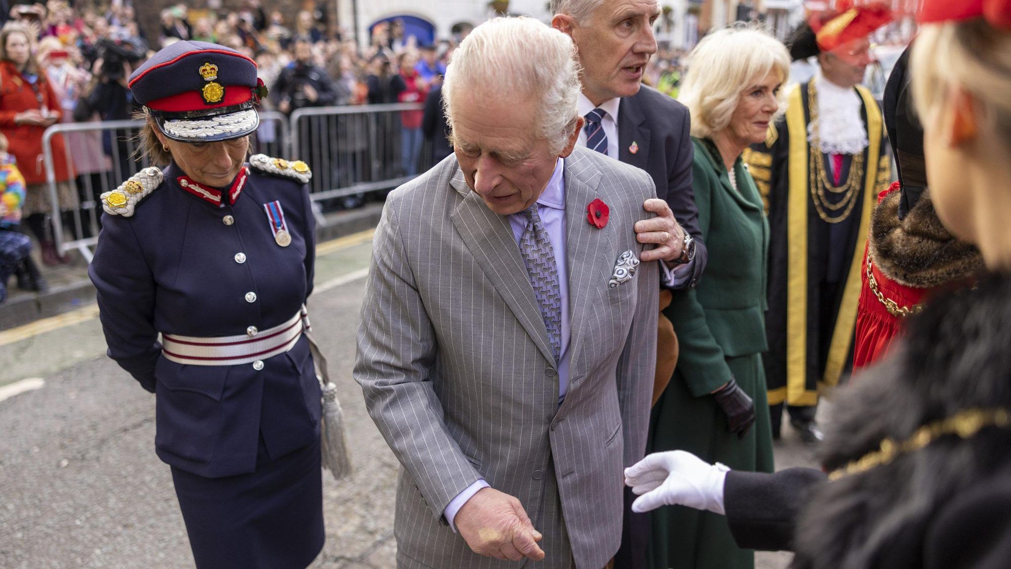 König Charles der Dritte schaut auf den Boden, nachdem ein Ei nach ihm geworfen wurde. Im Hintergrund ist eine Menschenmenge hinter einer Absperrung zu sehen. Neben Charles steht seine Gemahlin Camilla.