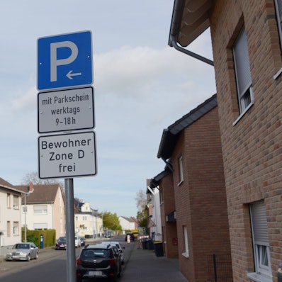 Ein Parken-Verkehrsschild steht auf einer Straße. Die Bewohner der Zone D können kostenfrei parken. Mehrere Autos stehen geparkt am Straßenrand.