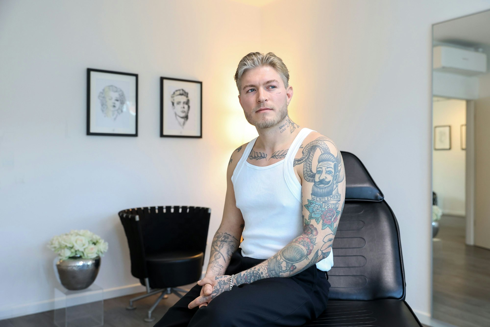 Das Bild zeigt Léon Sebastian Tiesler in einem weißen Unterhemd. Seine zahlreichen Tattoos auf dem Arm, am Brustbein, am Hals und im Gesicht sind gut zu erkennen.