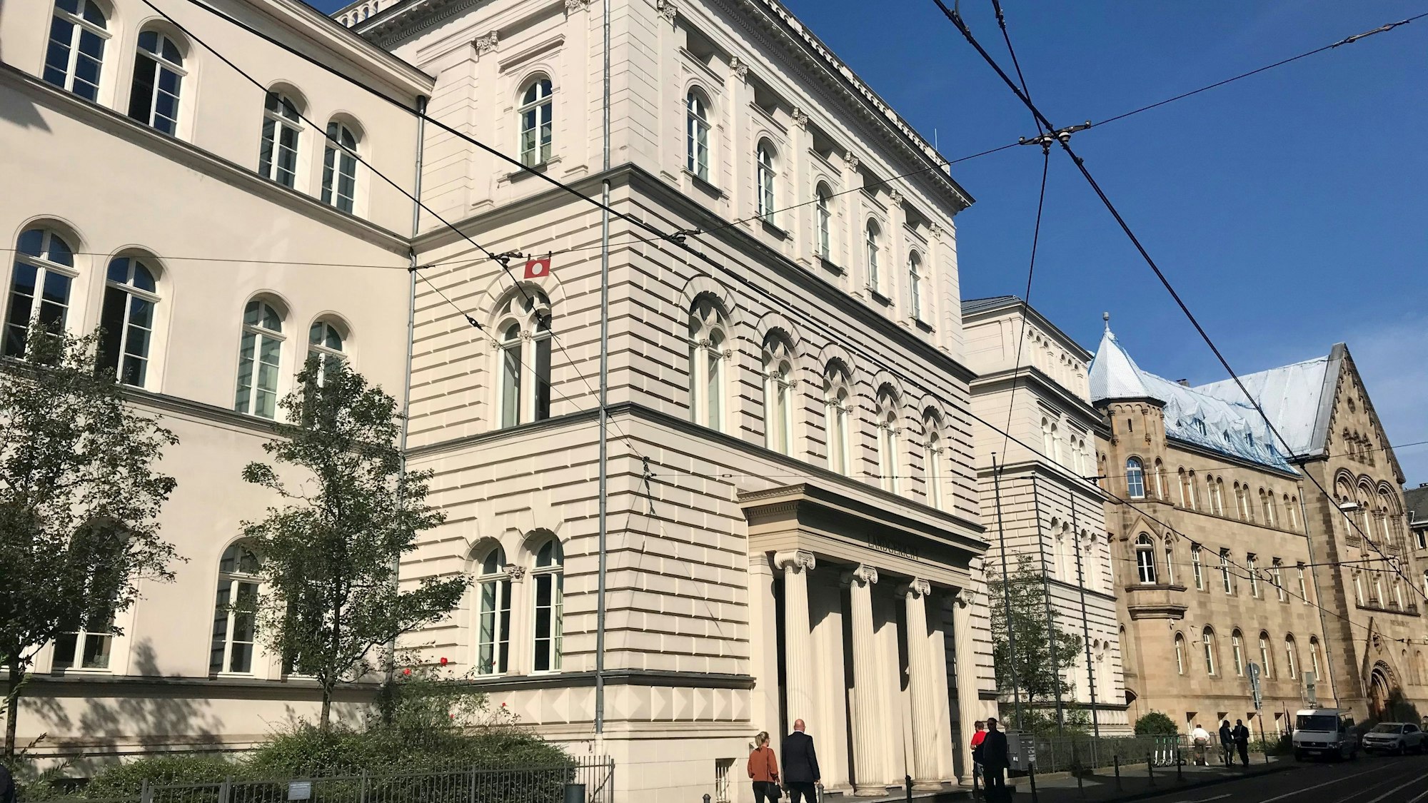 Ein großes, helles Gebäude mit einem Säulengang, auf der Straße gehen Passanten.