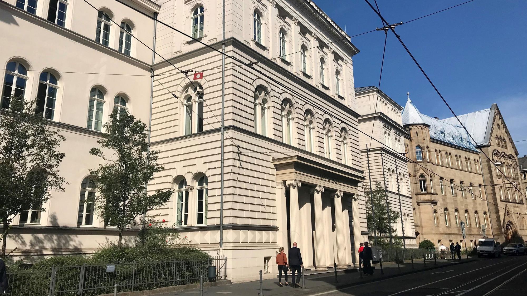 Ein großes altes Gebäude im Stil des Klassizismus und Säulen vor dem Eingang. Die Sonne scheint, der Himmel ist blau. LInks stehen Bäume, in der Straßen liegen Straßenbahnschienen.