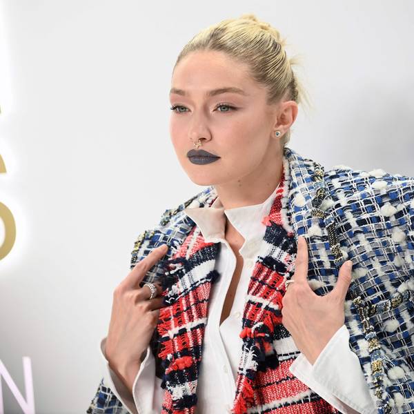 Gigi hadid steht vor hellem Hintergrund und schaut an der Kamera vorbei. Sie hält mit ihren Händen ihren Mantel über den Schultern fest.