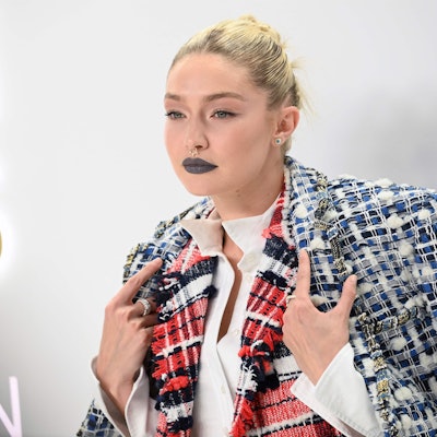 Gigi hadid steht vor hellem Hintergrund und schaut an der Kamera vorbei. Sie hält mit ihren Händen ihren Mantel über den Schultern fest.