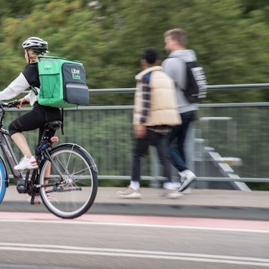 Eine Radfahrerin trägt einen grünen Rucksack von Uber Eats und fährt auf einem Radweg über eine Brücke.