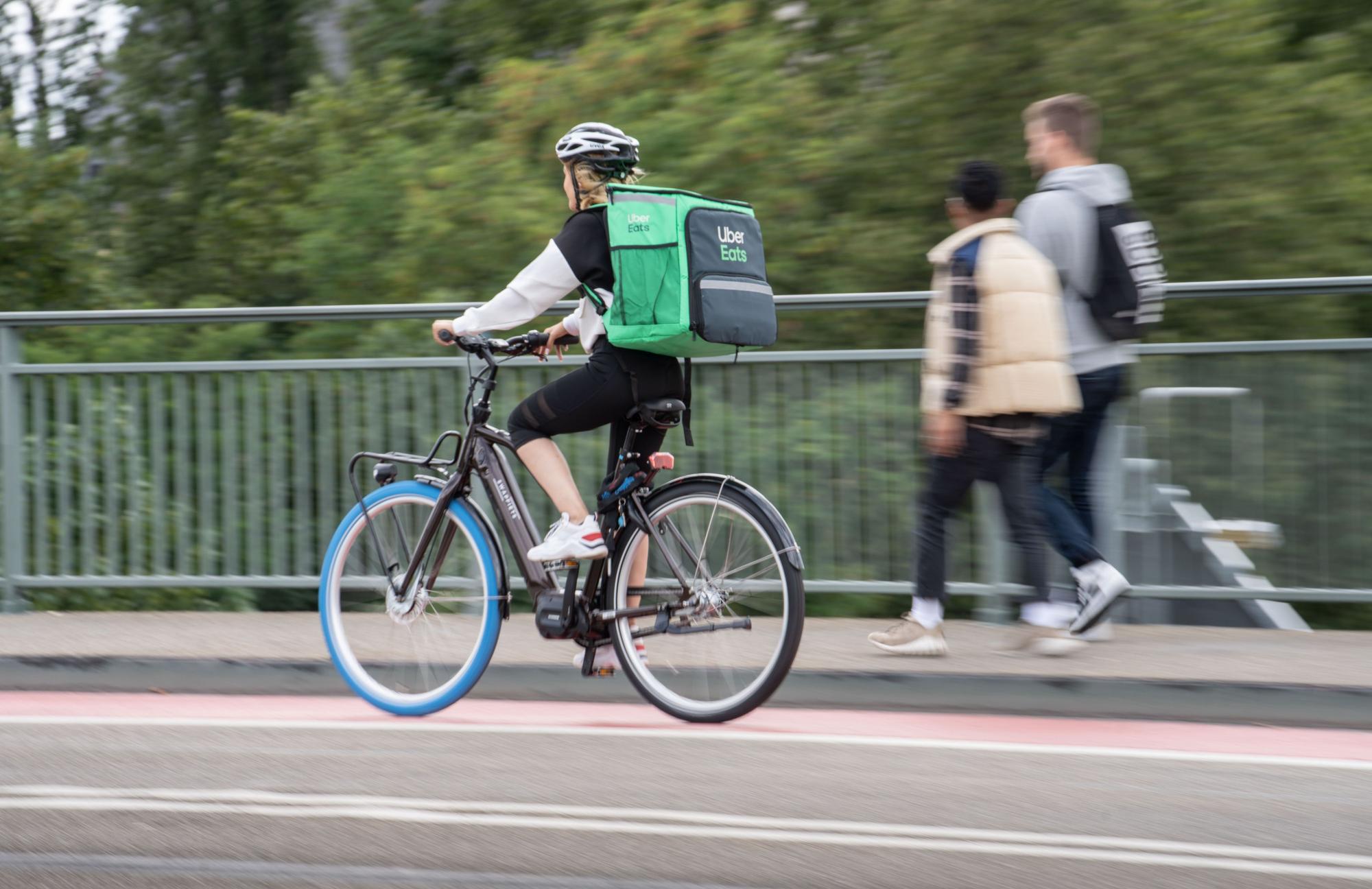 Eine Radfahrerin trägt einen grünen Rucksack von Uber Eats und fährt auf einem Radweg über eine Brücke.