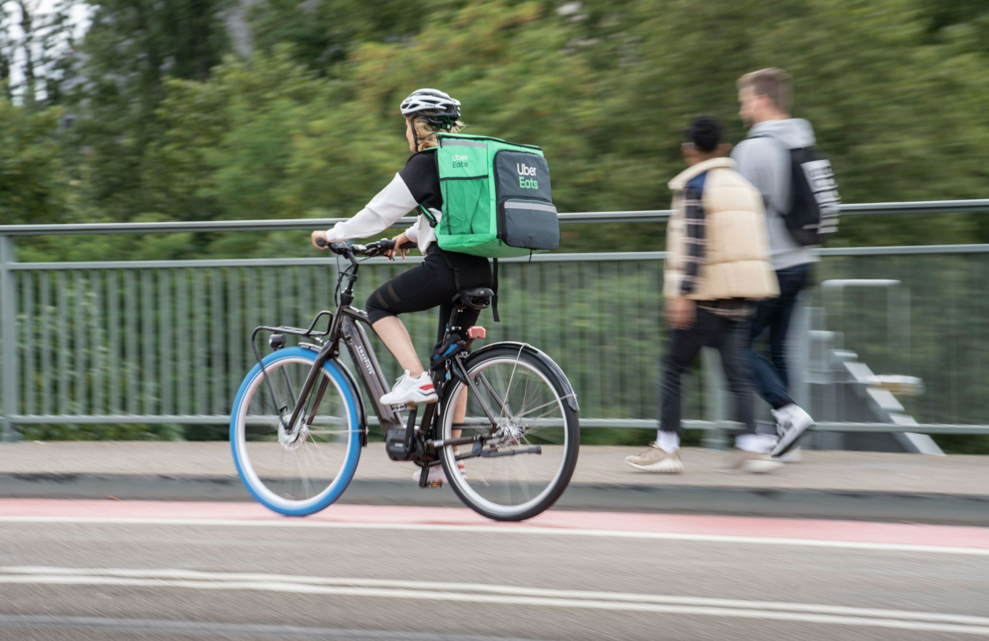 Eine Radfahrerin trägt einen grünen Rucksack von Uber Eats und fährt auf einem Radweg über eine Brücke.