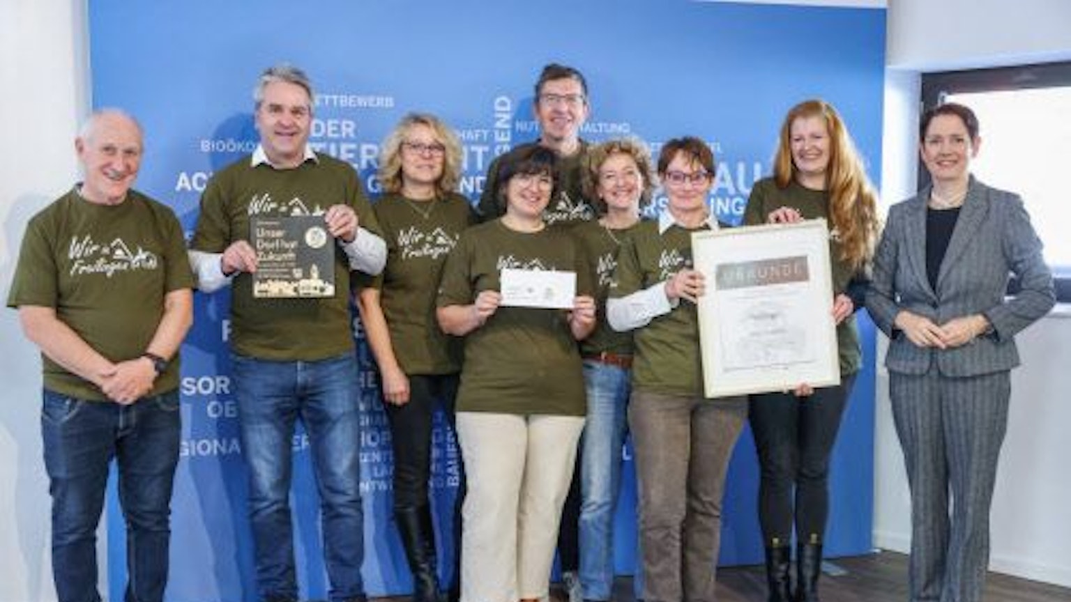 Eine Delegation von Preisträgern von „Unser Dorf hat Zukunft“ steht vor einer blauen Wand. Einige der Beteiligten halten Urkunden in die Kamera.