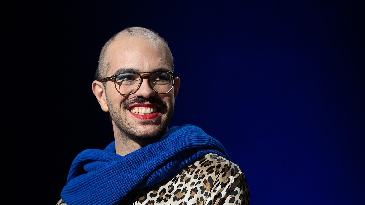 Kim de l'Horizon trägt blauen Schal und roten Lippenstift und lächelt in die Kamera.