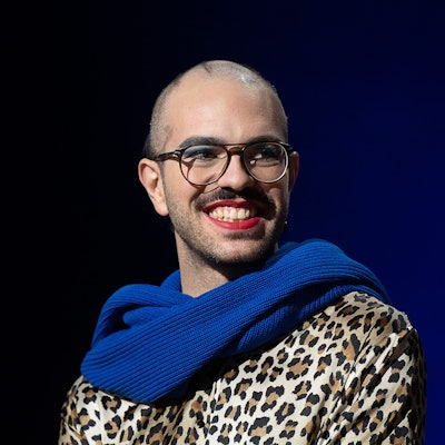 Kim de l'Horizon trägt blauen Schal und roten Lippenstift und lächelt in die Kamera.