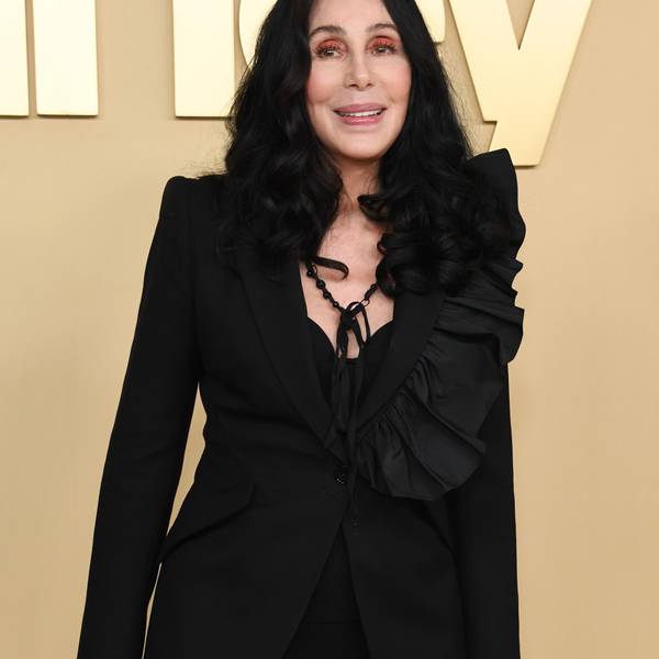 Cher lächelt für die Fotografen. Sie trägt ihre schwarzen Haare leicht gewellt und offen. Sie trägt eine schwarze Bluse und eine schwarze Kette.