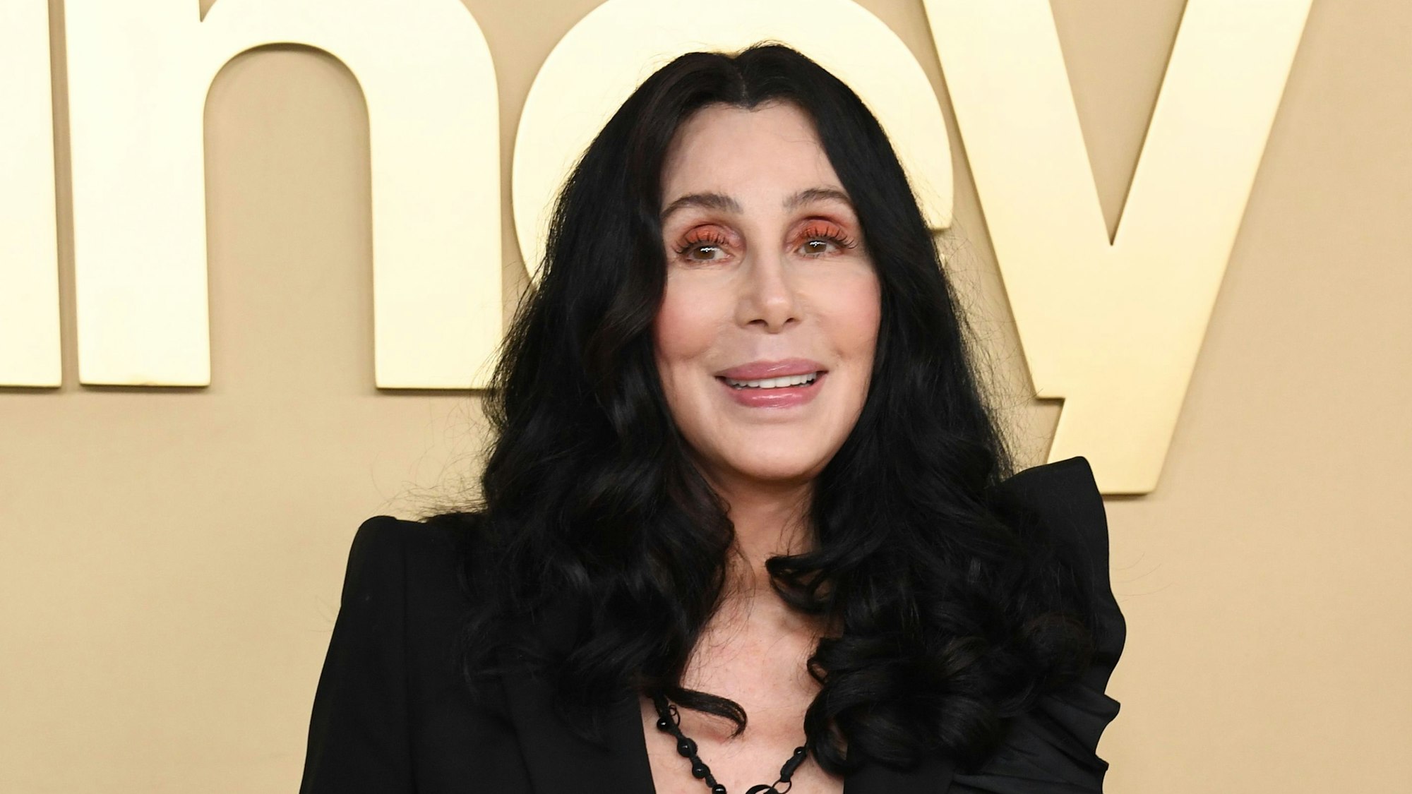 Cher lächelt für die Fotografen. Sie trägt ihre schwarzen Haare leicht gewellt und offen. Sie trägt eine schwarze Bluse und eine schwarze Kette.