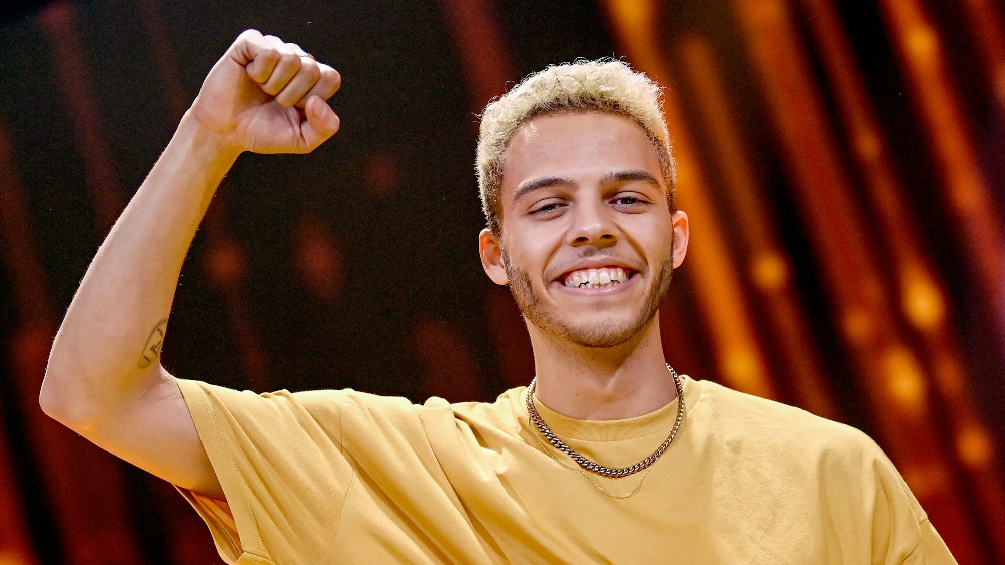 Malik Harris freut sich bei der Show „Germany 12 Points - der deutsche ESC-Vorentscheid“ über den Sieg. Er ballt die Faust und streckt den Arm nach oben. Er trägt eine Goldkette und ein gelbes T-Shirt.
