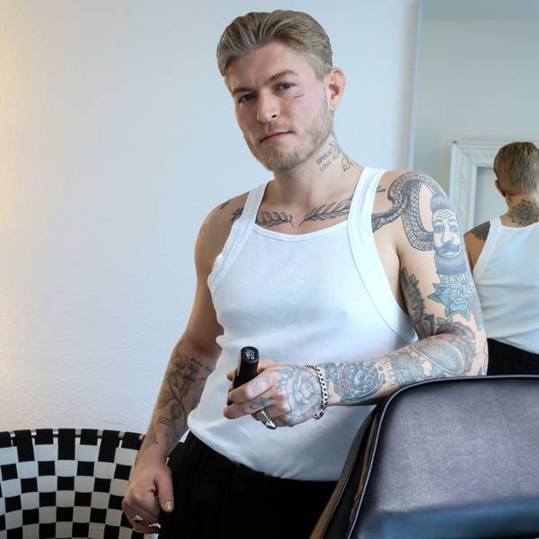 Das Bild zeigt Léon Sebastian Tiesler in einem weißen Unterhemd. Seine zahlreichen Tattoos auf dem Arm, am Brustbein, am Hals und im Gesicht sind gut zu erkennen.